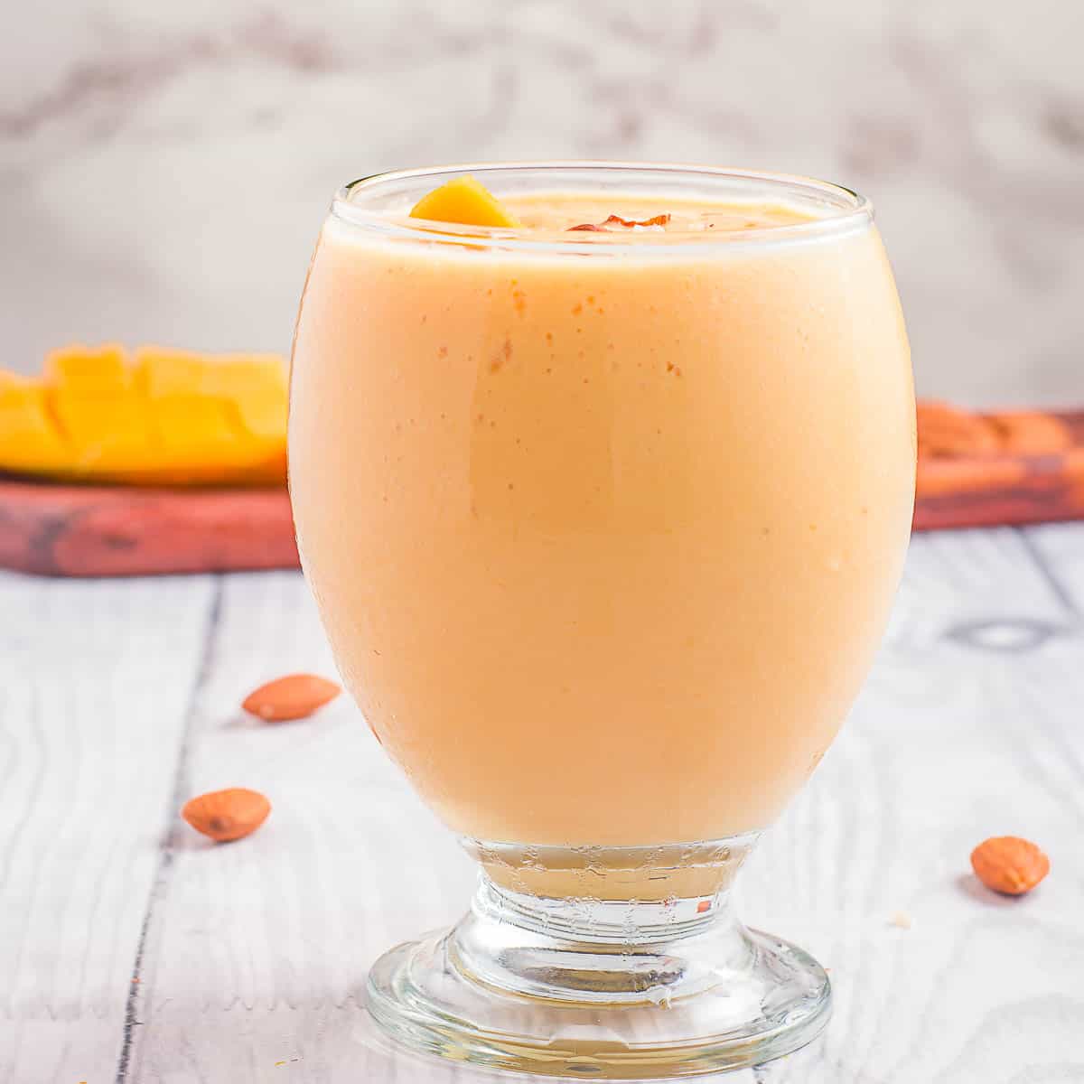 Side shot of mango lassi in a clear curvy glass garnished with mango chunks and pieces of almonds with more almonds and mango in the background.