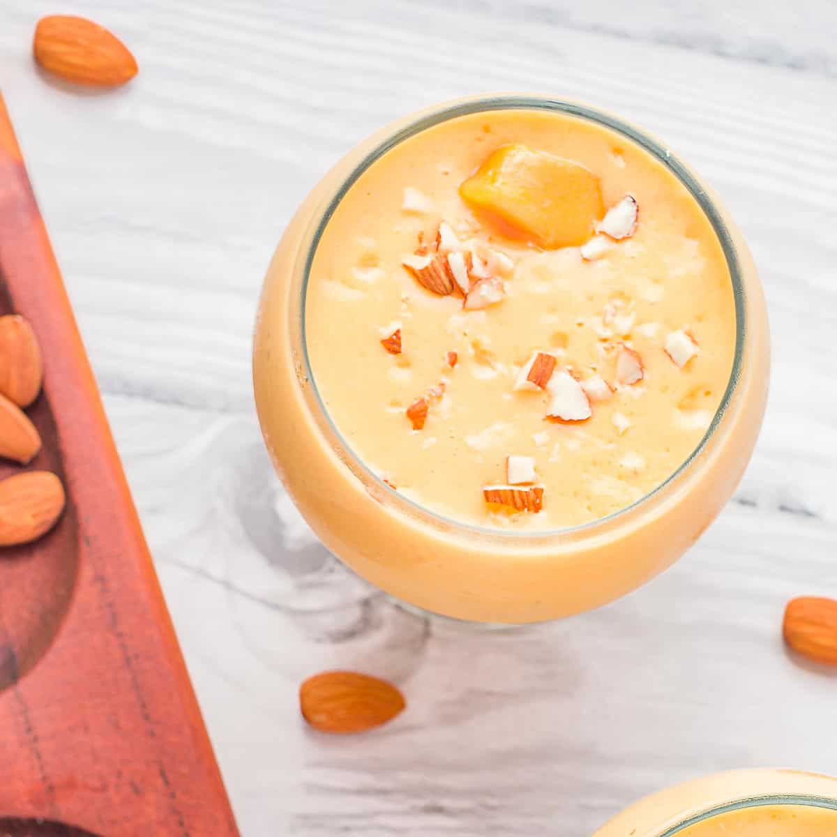 Overhead shot of mango lassi in a clear curvy glass garnished with mango chunks and pieces of almonds with more almonds in the background.