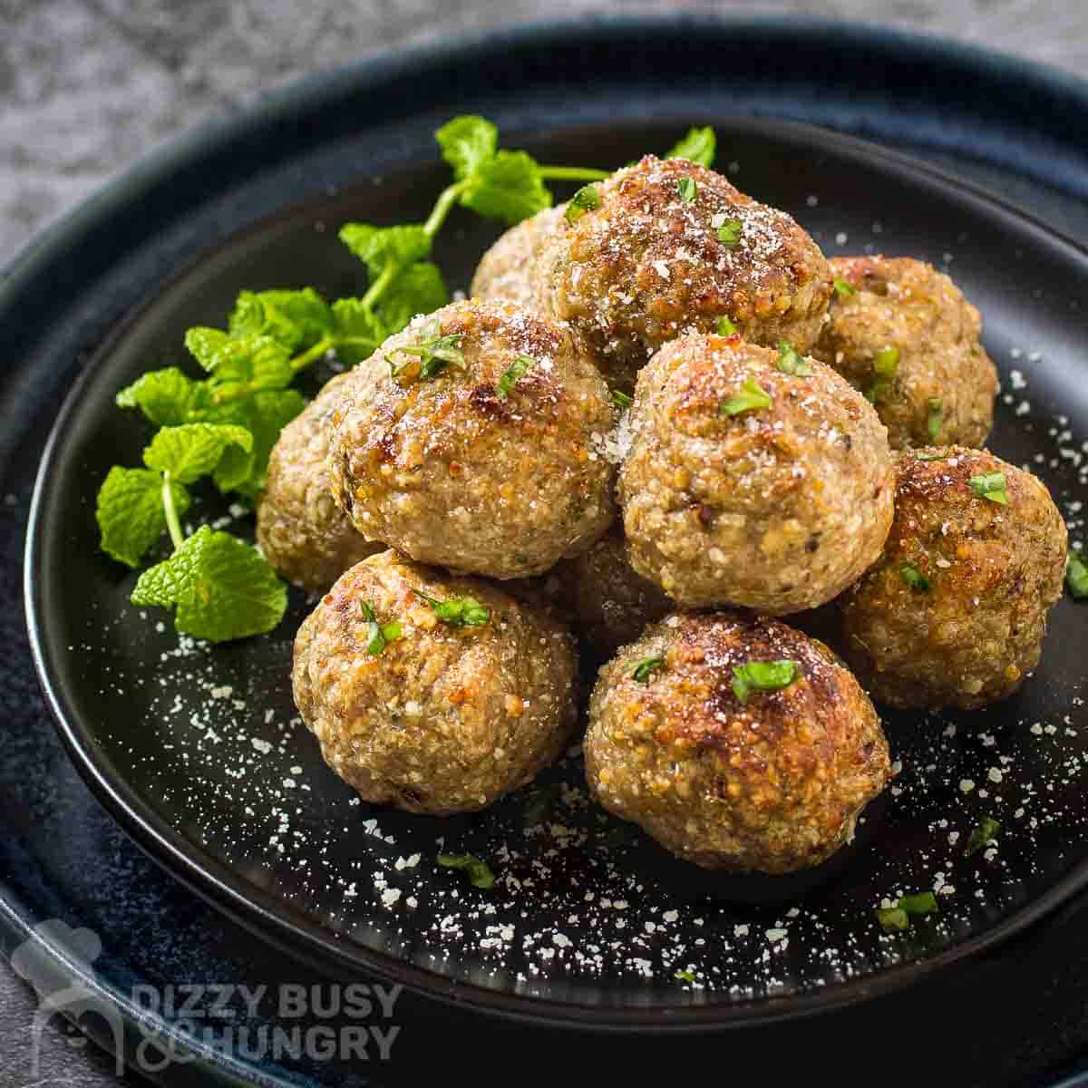 Side shot of multiple meatballs stacked on a black plate garnished with herbs and parmesan cheese.