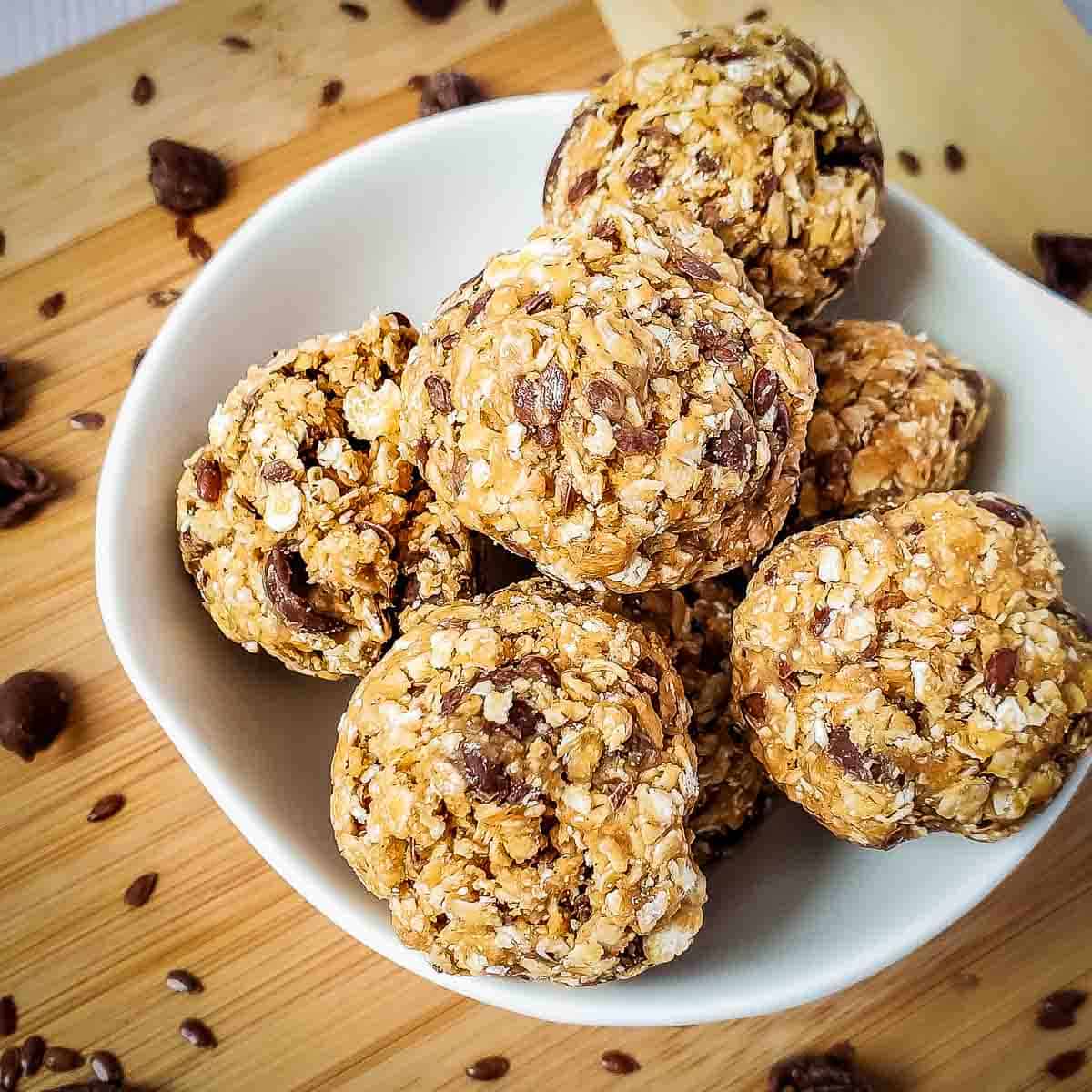 Side shot of multiple energy balls in a white bowl on a cutting board sprinkled with flax seeds and chocolate chips.