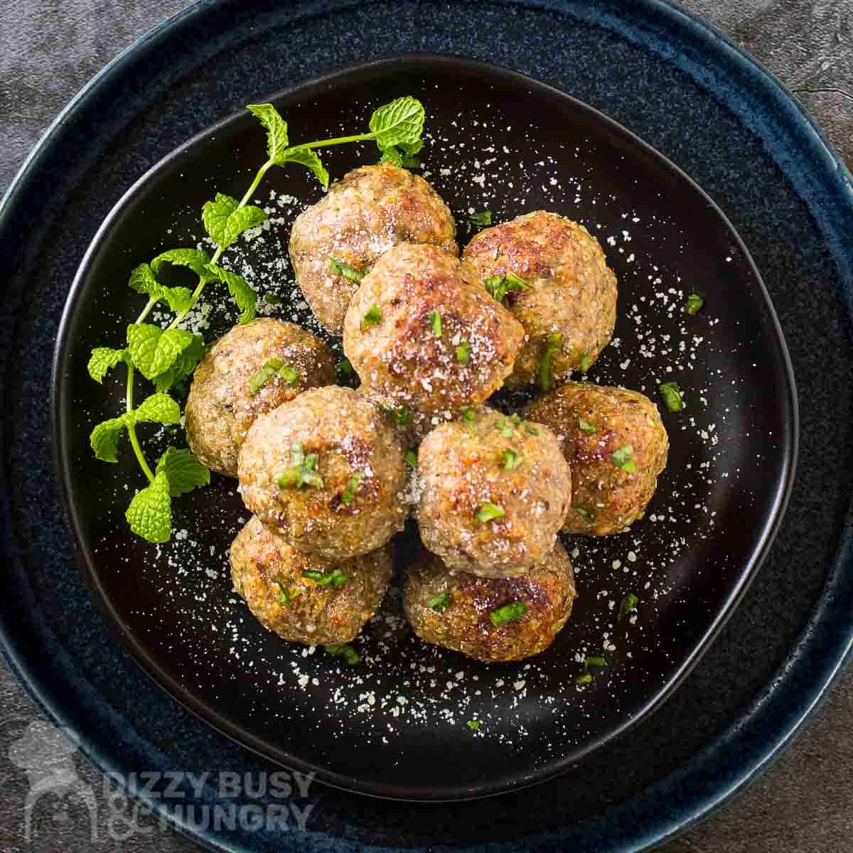 Overhead shot of multiple meatballs stacked on a black plate garnished with herbs and parmesan cheese.