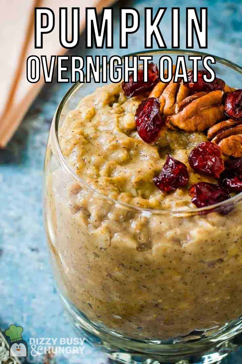 Close up shot of overnight oats in a clear glass cup garnished with pecans and cranberries on a blue speckled surface with peach colored napkins in the background.