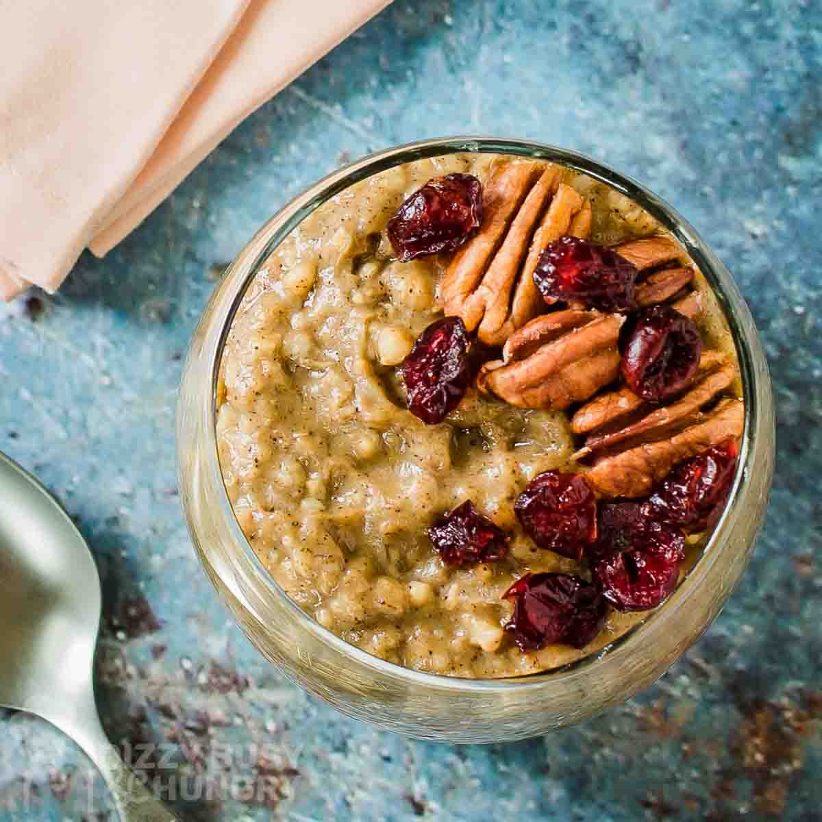Overhead shot of overnight oats in a clear glass cup garnished with pecans and cranberries on a blue speckled surface with a spoon and peach colored napkins in the background.