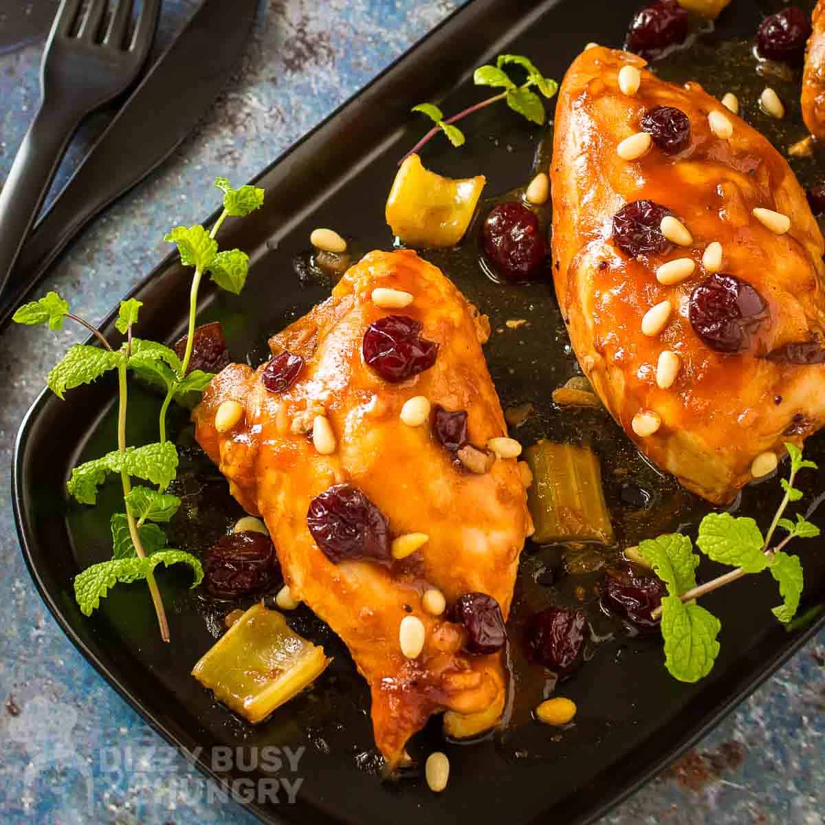 Overhead shot of three pieces of cranberry chicken garnished with cranberries and herbs on a black plate with cutlery on the side.