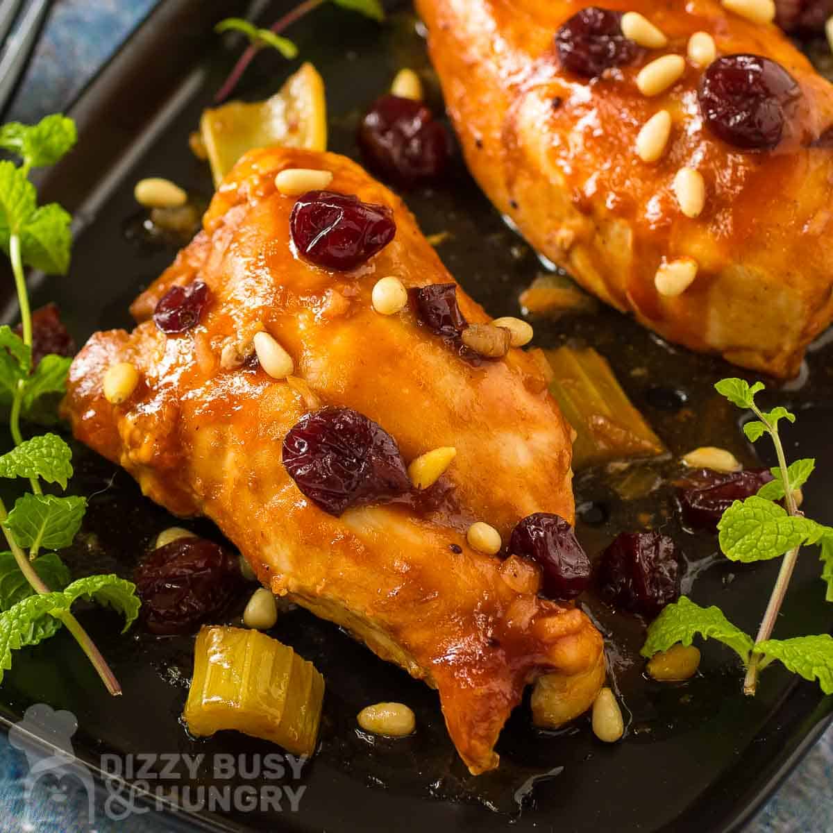 Overhead close up shot of two pieces of cranberry chicken garnished with cranberries and herbs on a black plate.