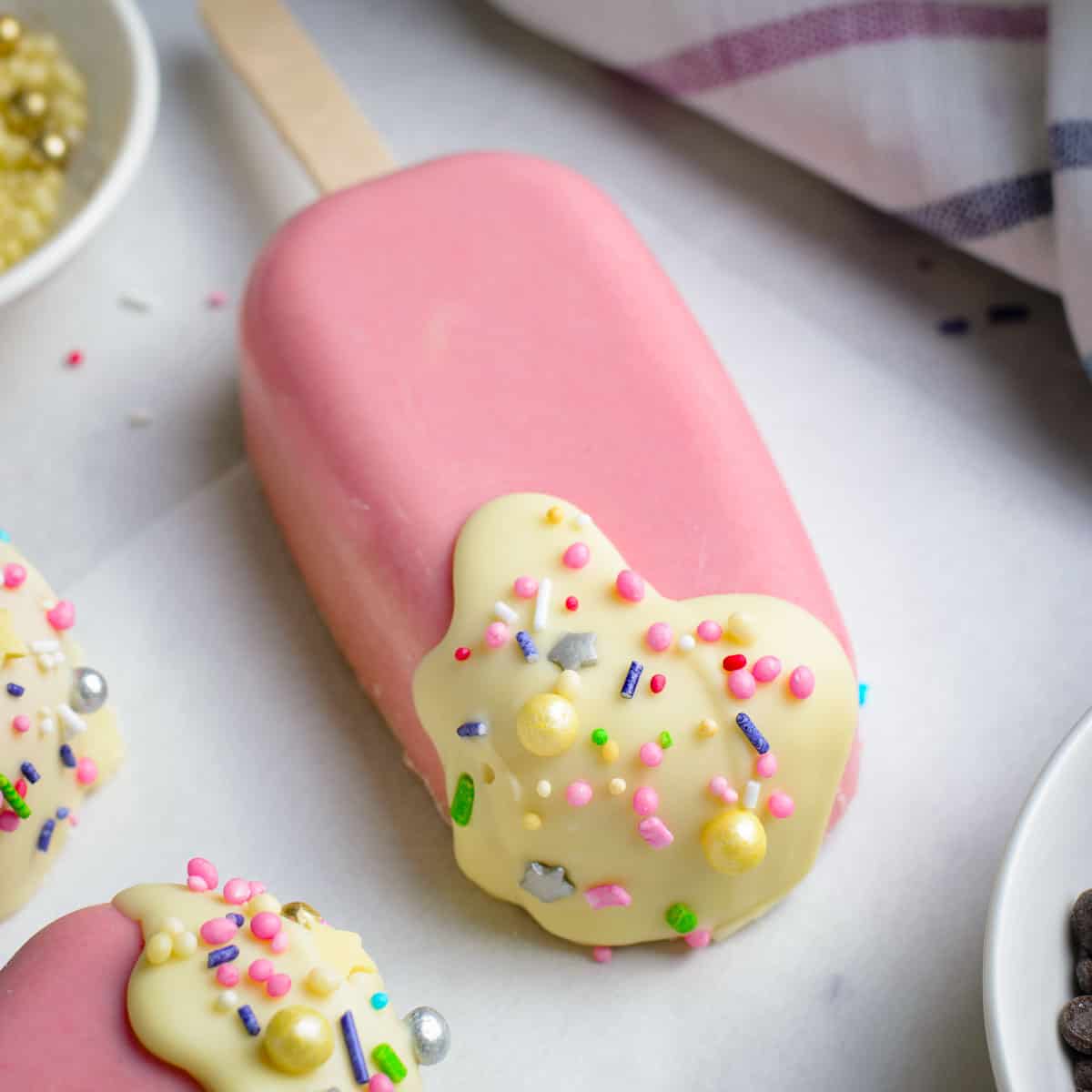 Close up shot of a brownie cakesicle garnished with sprinkles on a white surface with more sprinkles in the background.