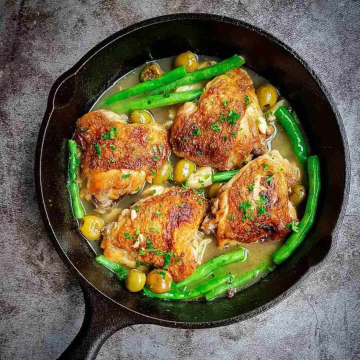 Overhead shot of pan fried chicken thighs with olives and green beans in a skillet on a black surface.