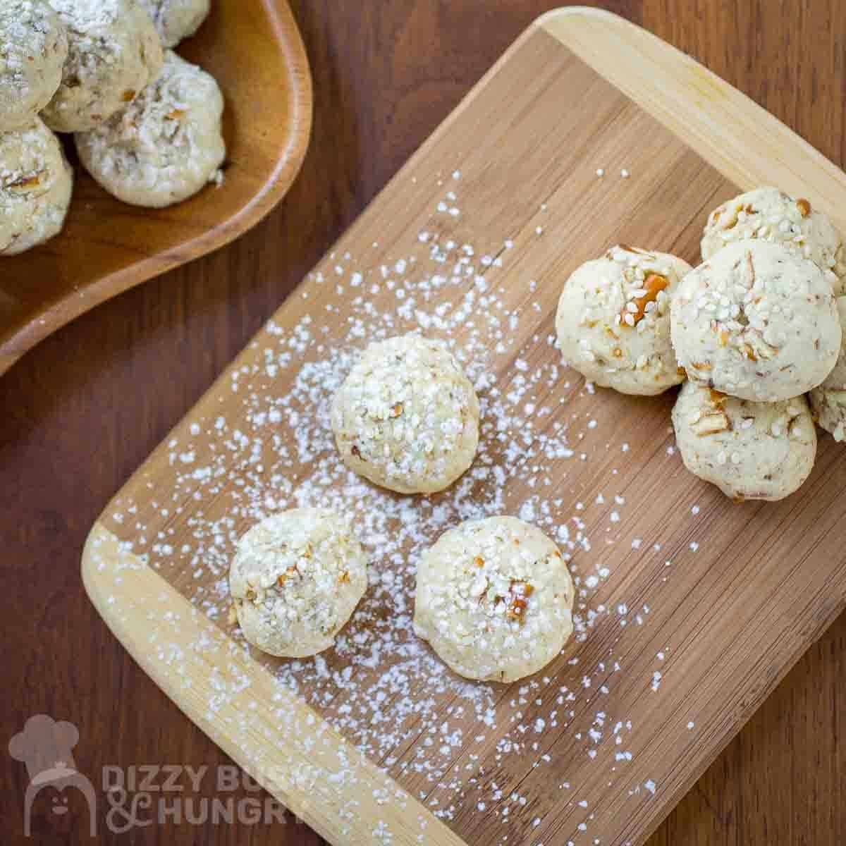 Overhead shot of multiple sesame cookies on a wooden cutting board garnished with powdered sugar with more cookies in the background.