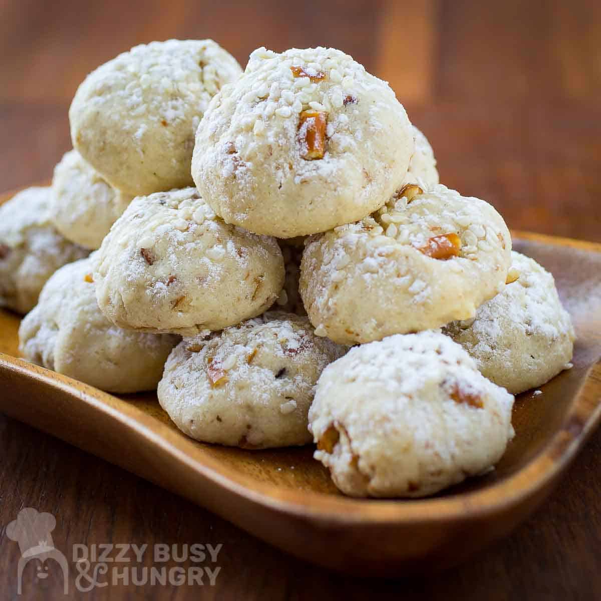 Side view of multiple sesame cookies stacked on a wooden bowl-plate garnished with powdered sugar.