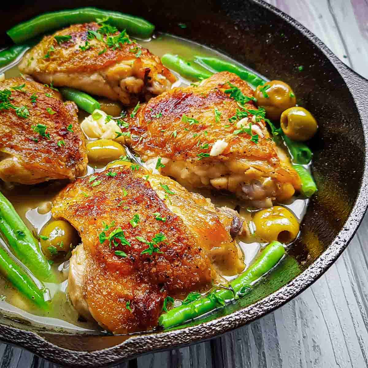 Side close up shot of pan fried chicken thighs with olives and green beans in a skillet on a black surface.
