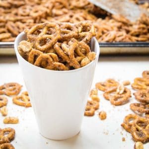 Side angled view of cinnamon pretzels in a white ceramic cup with more pretzels in the background on a baking sheet and scattered on a white surface.