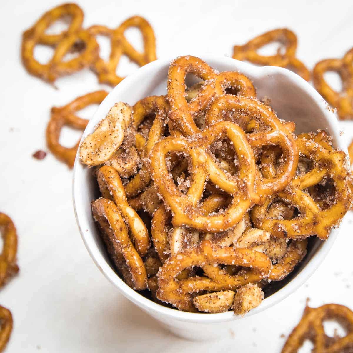 Overhead shot of cinnamon pretzels in a white cup with more scattered on a white surface in the background.