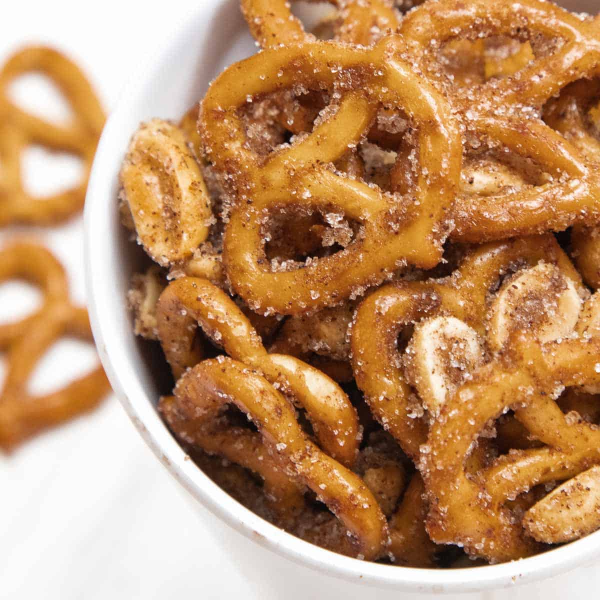 Overhead close up shot of cinnamon pretzels in a white cup with more scattered on a white surface in the background.