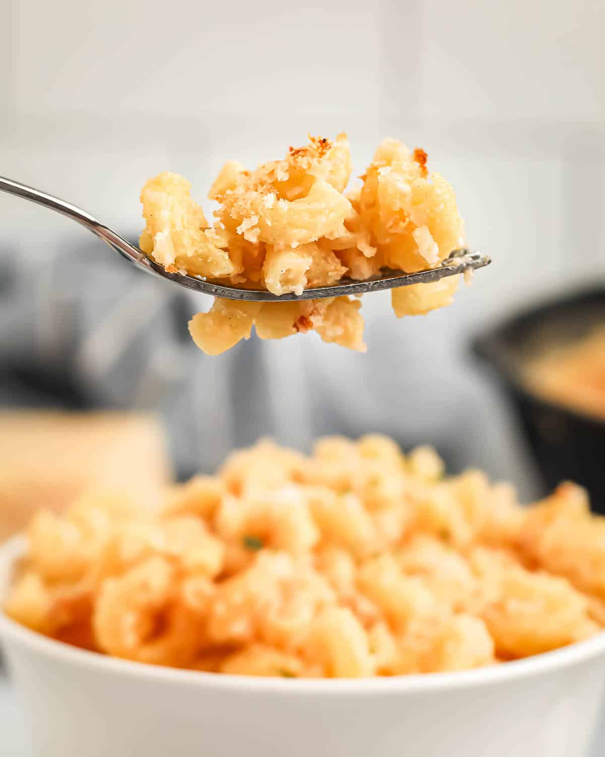 Side shot of a fork holding a bite of Mac and cheese with a white bowl of Mac and cheese in the background.
