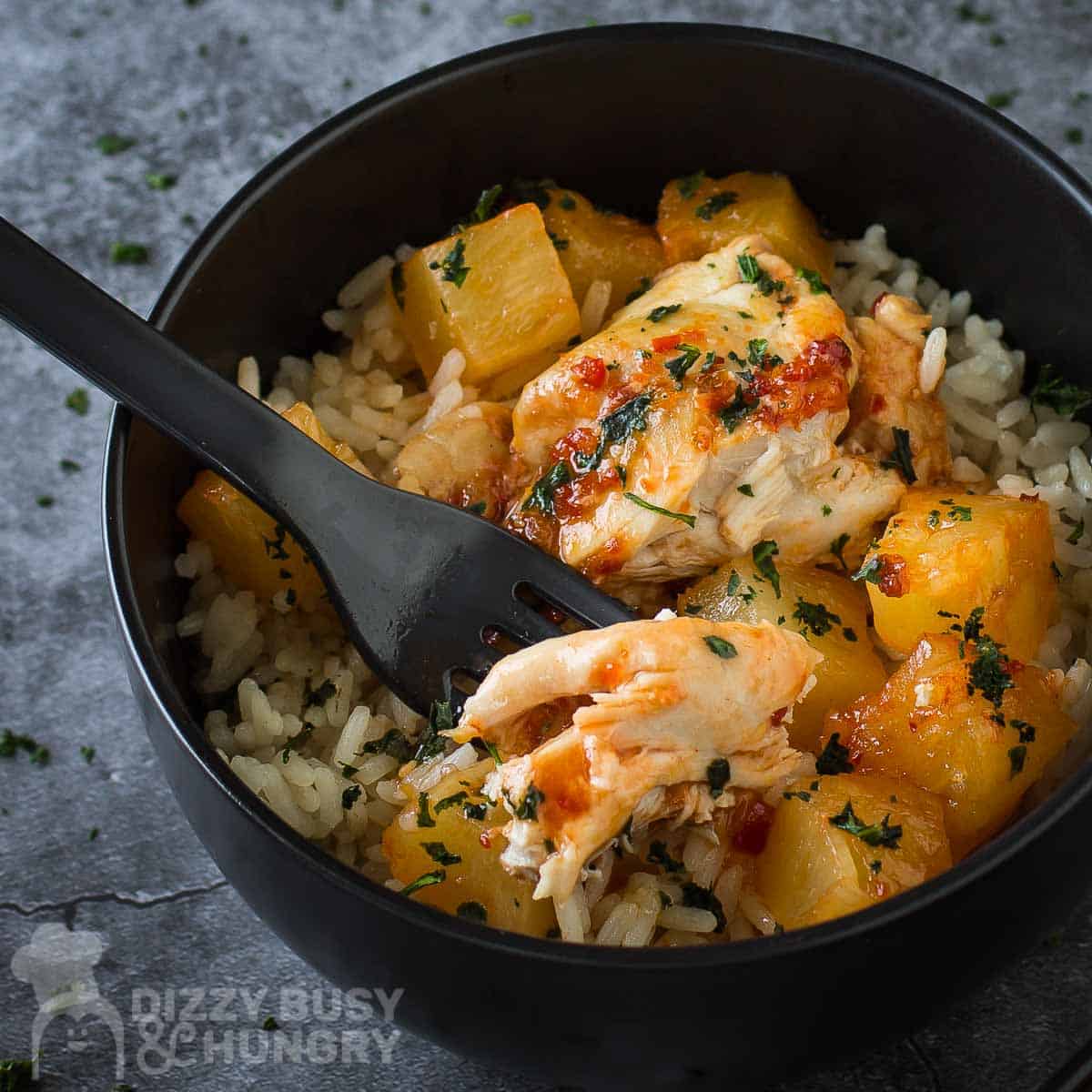 Side close up shot of Hawaiian chicken in a black bowl with rice and a black fork on the side on a grey marble countertop.