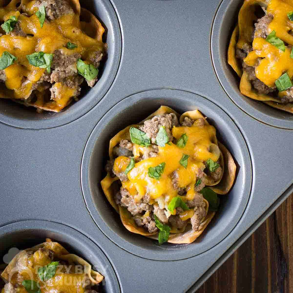 Overhead shot of multiple wonton cheeseburger cups in a cupcake sheet on a wooden surface.