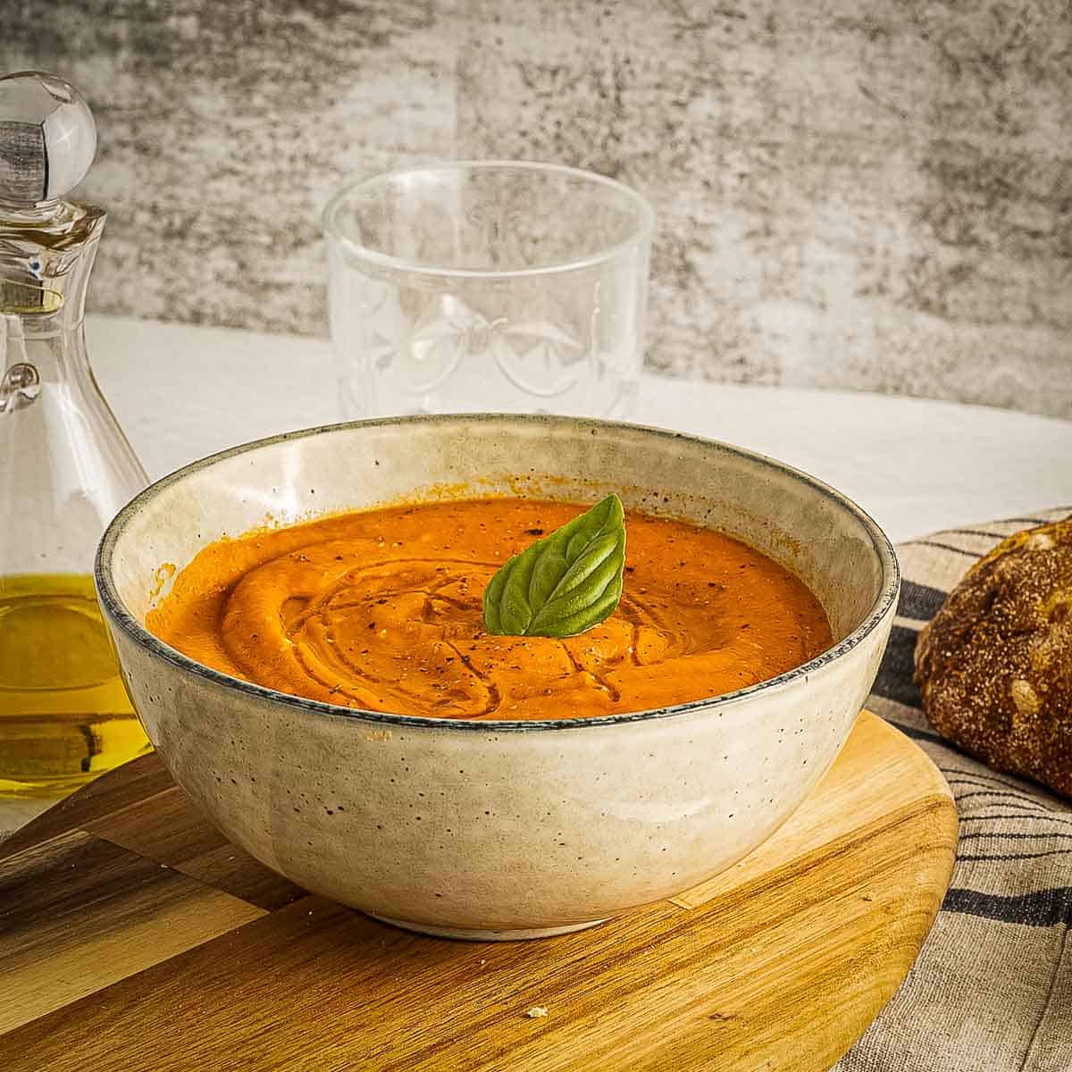 Side shot of tomato carrot soup in a tan bowl on a cutting board with a glass jar of olive oil, a drinking glass, and a loaf of bread in the background.