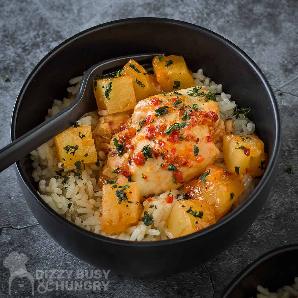 Side close up shot of Hawaiian chicken in a black bowl with rice and a black fork on the side on a grey marble countertop.