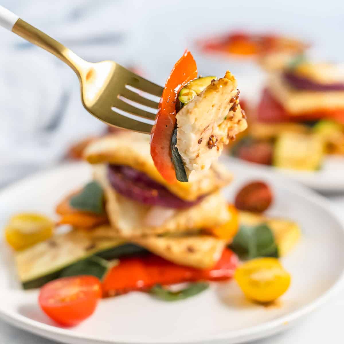 Side close up view of a fork holding up a bite of colorful halloumi salad with two more white plates of it on a white surface with cherry tomatoes in the background.