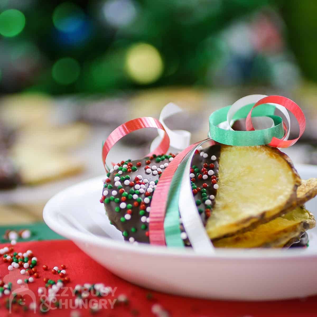 Side shot of multiple chocolate covered potato chips wrapped in red, green, and white ribbon on a small white plate on red and green tissue paper.