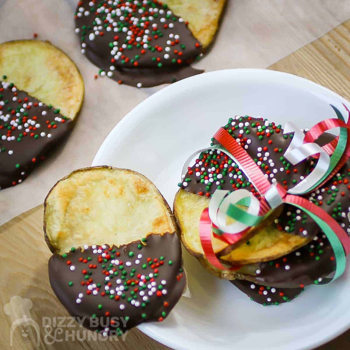 Overhead close up shot of multiple chocolate covered potato chips wrapped in Christmas ribbons on a white plate with more in the background on a wooden surface.