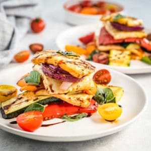 Side view of two white plates of colorful halloumi salad garnished with herbs on a white surface with cherry tomatoes in the background.