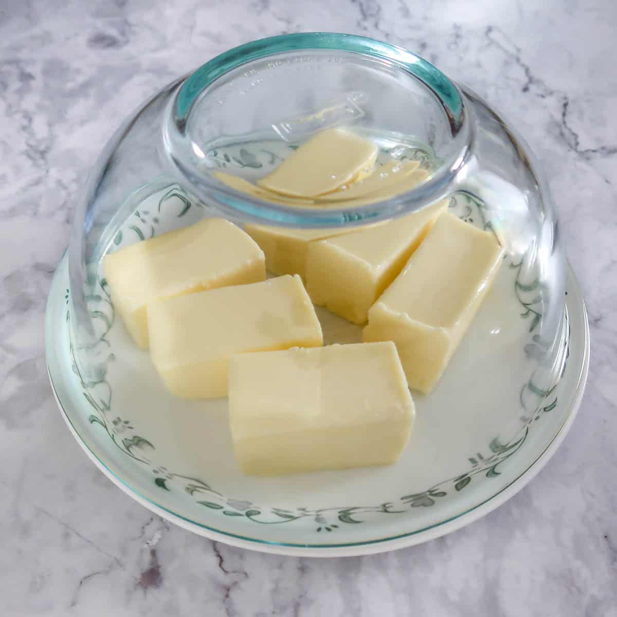 Side view of six half sticks of butter on a white plate with a clear bowl on top all on a white marbled table.