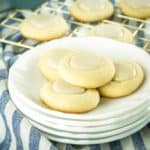 Side shot of four maple thumbprint cookies on a stack of white plates on a blue and white striped cloth and more cookies in the background on a gold drying rack.