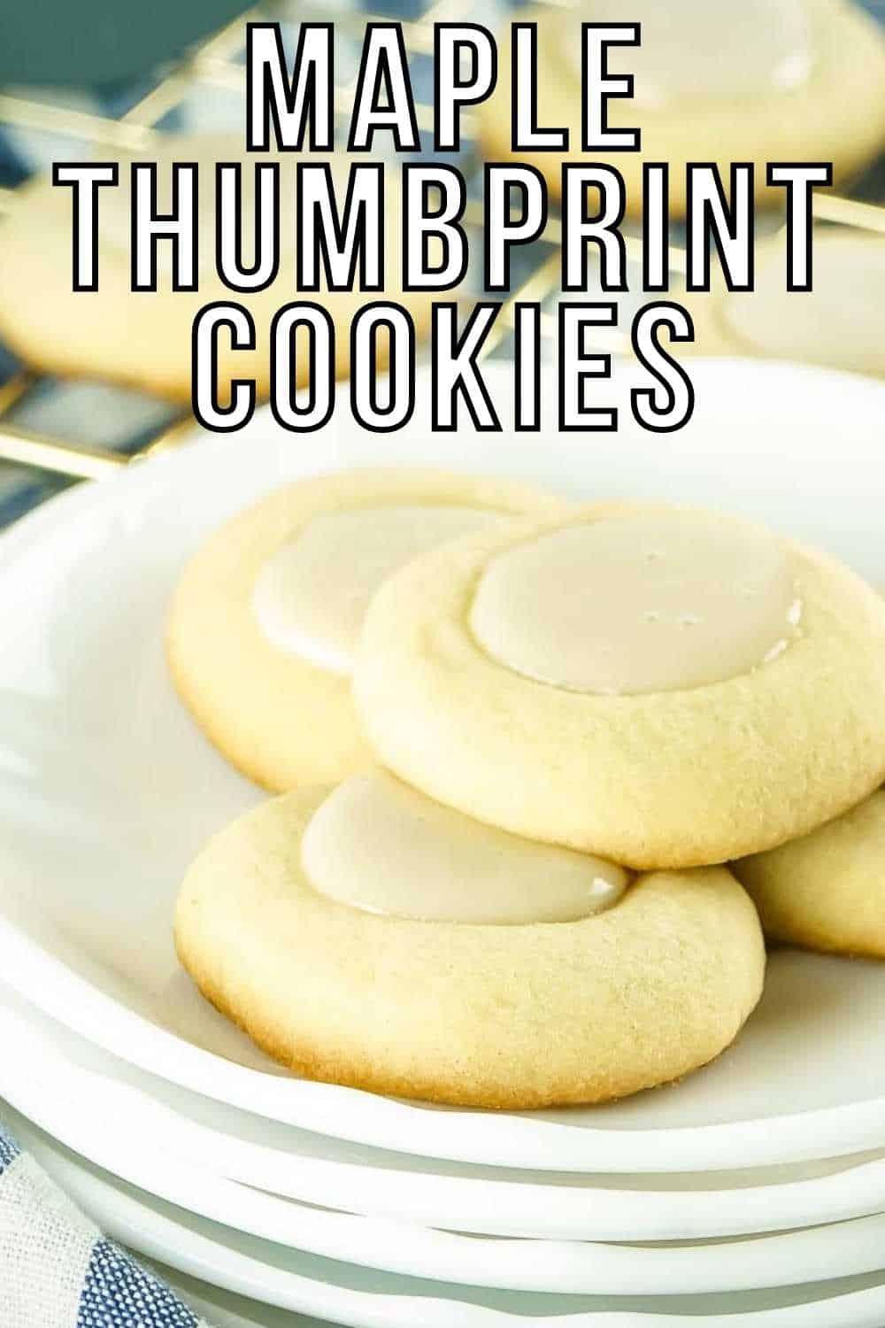 Side close up shot of four maple thumbprint cookies on a stack of white plates on a blue and white striped cloth and more cookies in the background on a gold drying rack.