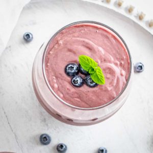 Overhead shot of acai smoothie garnished with blueberries and mint in a clear glass on a white surface with blueberries scattered around.