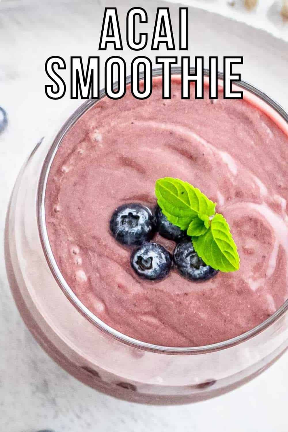 Overhead close up shot of acai smoothie garnished with blueberries and mint in a clear glass on a white surface.