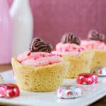 Side shot of three cookie cups in a row on a white plate with chocolate hearts scattered around with a pink background.