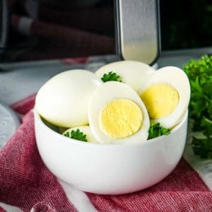 Side shot of multiple boiled eggs, some halved, with some herbs on a white bowl on a red tablecloth.