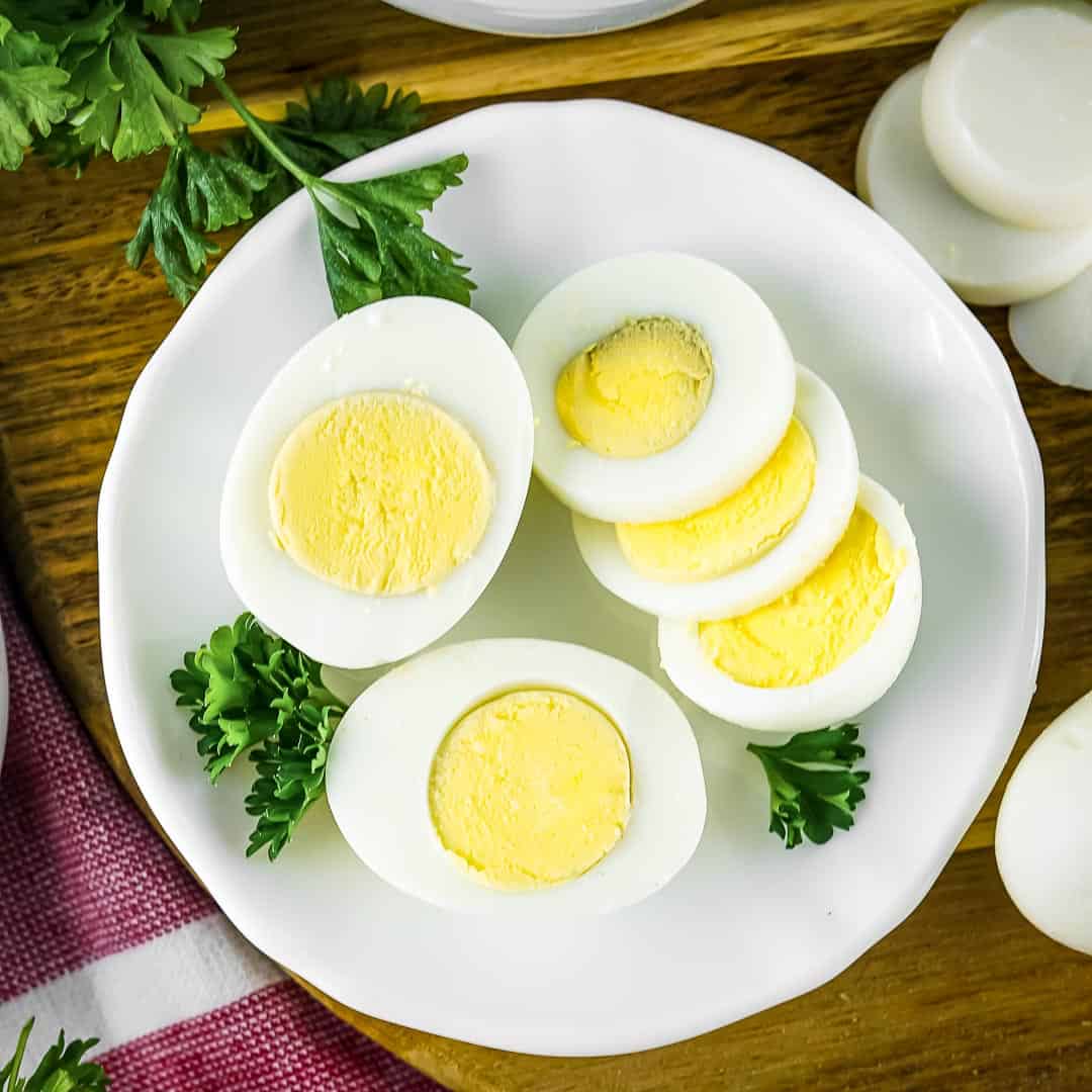 Overhead shot of a few sliced boiled eggs garnished with herbs on a white plate on a wooden surface with a red tablecloth on the side.