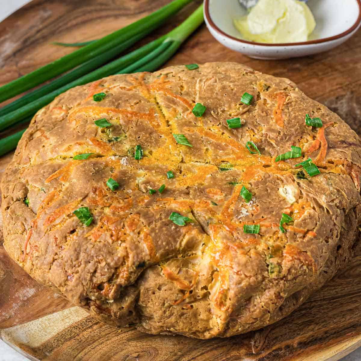 Side shot of whole loaf of cheddar bread garnished with chives on a wooden cutting board with whole chives and a plate of butter in the background.