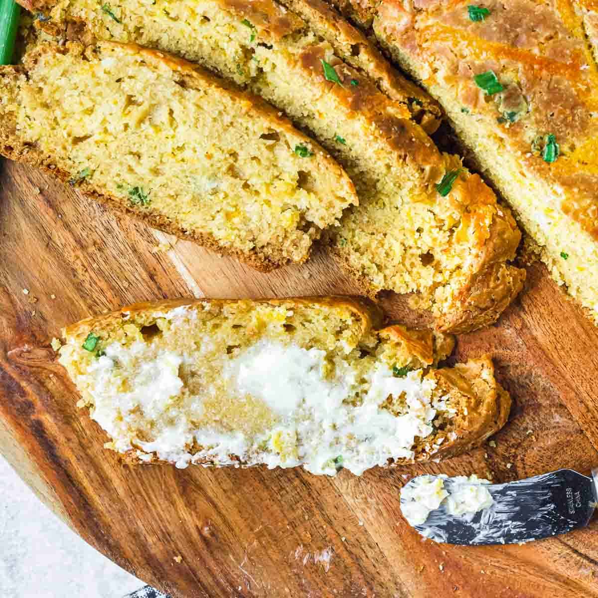 Overhead shot of multiple slices of cheddar bread, one with butter spread on it, on a wooden cutting board with a butter knife on the side.