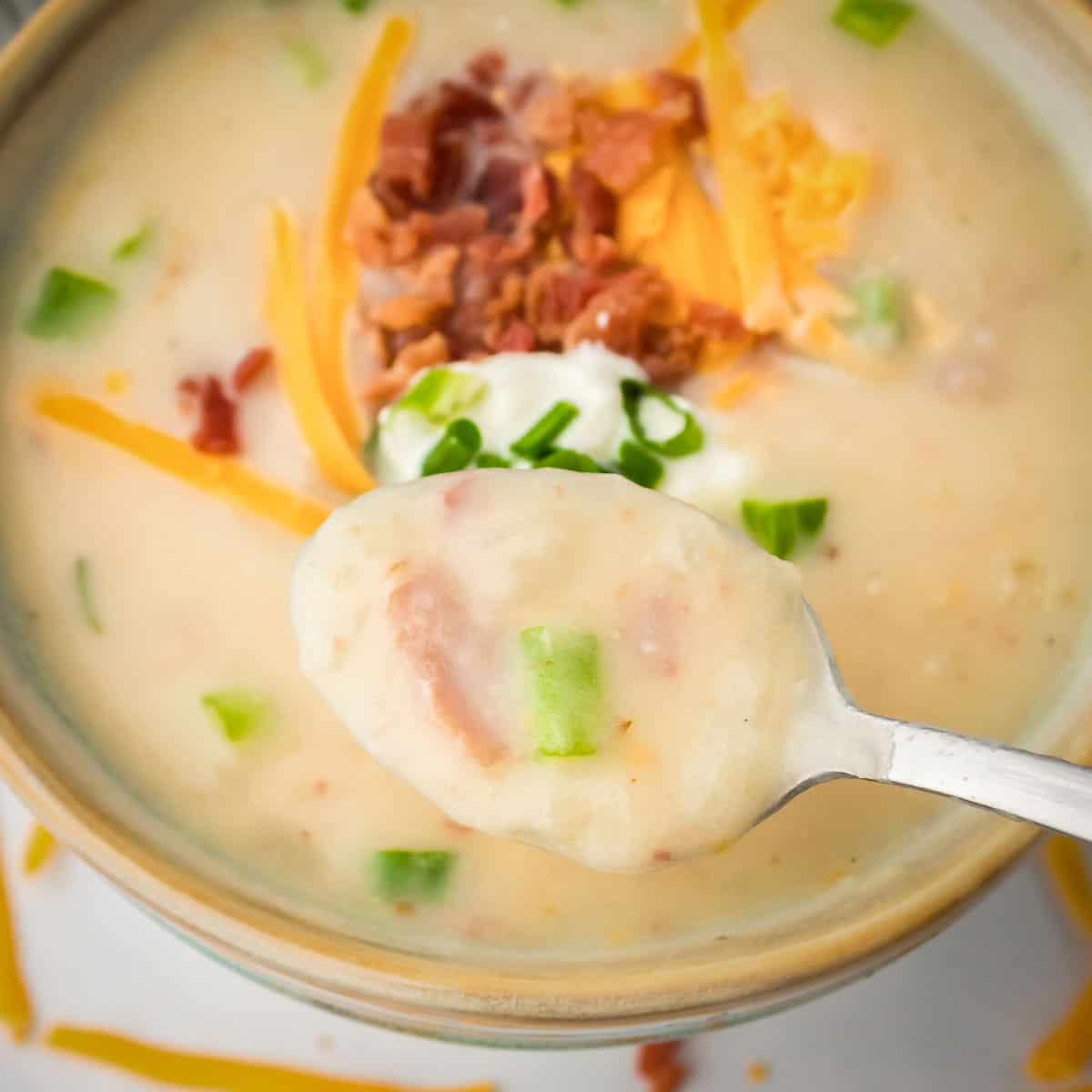 Overhead shot of a spoon holding some potato soup garnished with bacon, cheese, and chives in a green bowl.