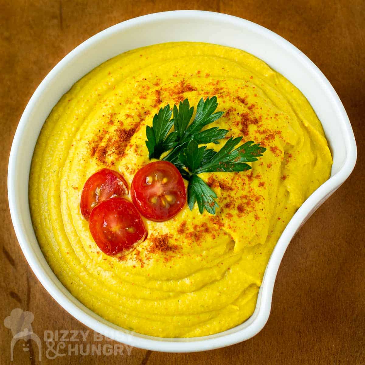 Overhead view of hummus garnished with halved cherry tomatoes, herbs, and paprika in a white bowl on a wooden surface.