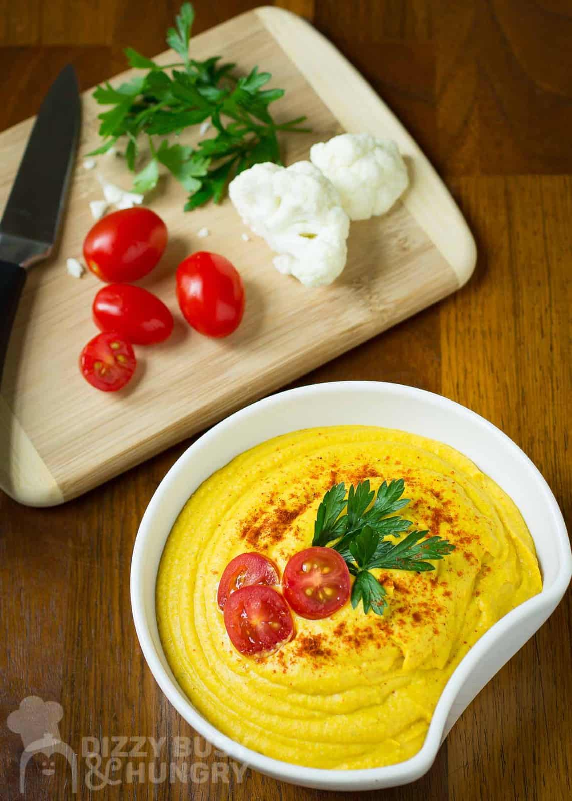 Side shot of hummus in a white bowl garnished with cherry tomatoes, herbs, and paprika with a wooden cutting board with cherry tomatoes, cauliflower, herbs, and a knife in the background all on a wooden surface.