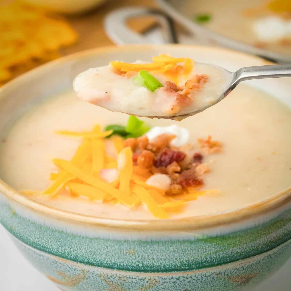 Side close up shot of a spoon holding some potato soup garnished with bacon, cheese, and chives in a green bowl.