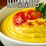 Close up side shot of hummus garnished with halved cherry tomatoes, herbs, and paprika in a white bowl on a wooden surface.