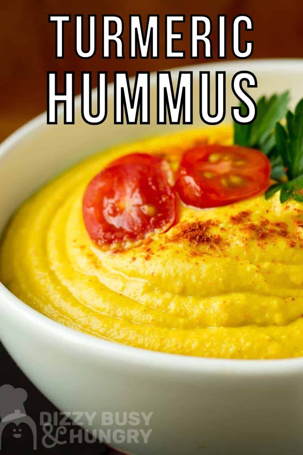 Close up side shot of hummus garnished with halved cherry tomatoes, herbs, and paprika in a white bowl on a wooden surface.