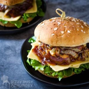 Side shot of barbecue burger with lettuce, cheese, caramelized onions, and a sesame bun on a black plate on a blue surface.