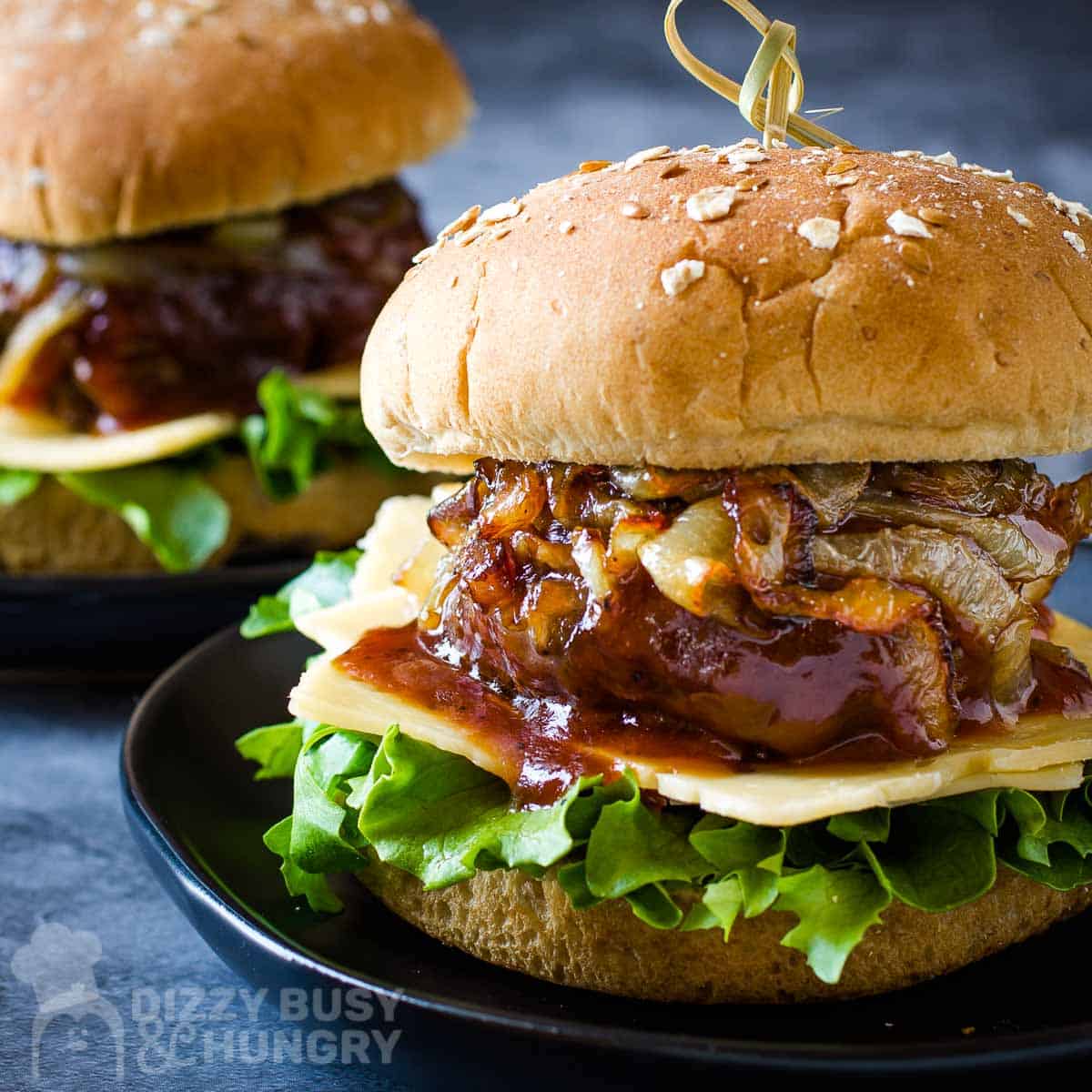 Side shot of two barbecue burgers with lettuce, cheese, caramelized onions, and a sesame bun on a black plate on a blue surface.