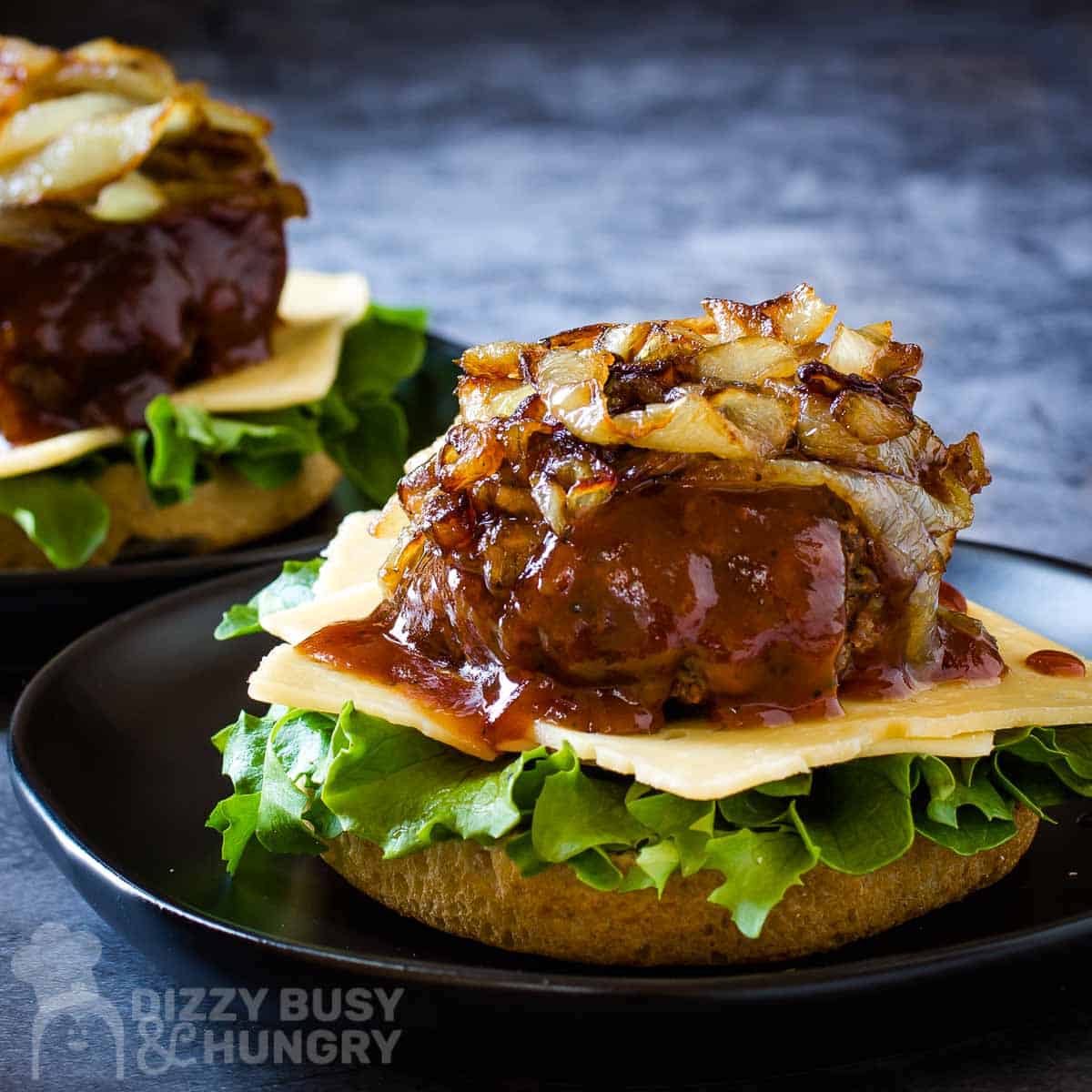 Side shot of two barbecue burgers with lettuce, cheese, and caramelized onions without a top bun on a black plate on a blue surface.