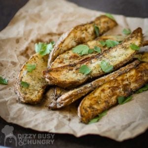 Side close up shot of multiple dill French fries garnished with herbs on a brown paper on a black surface.