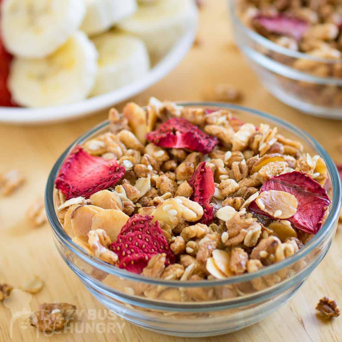 Side view of granola and strawberries in a clear bowl on a wooden surface with more granola and sliced fruits in the background.