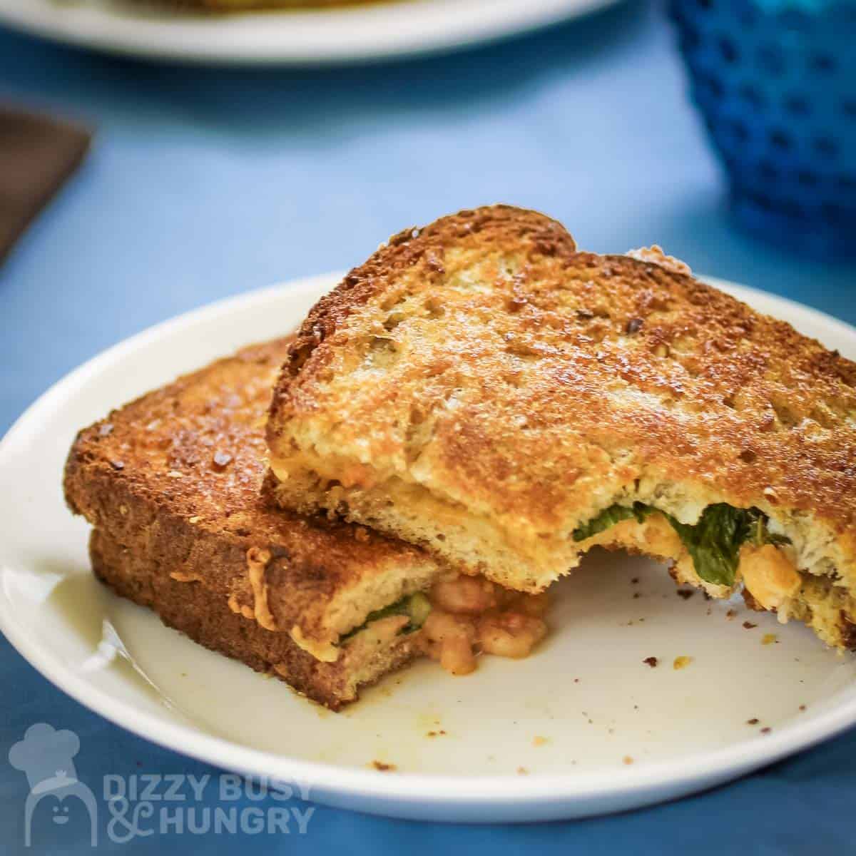 Side shot of two halves of grilled cheese layered on a white plate, the top on with a bite taken out, all on a blue surface.