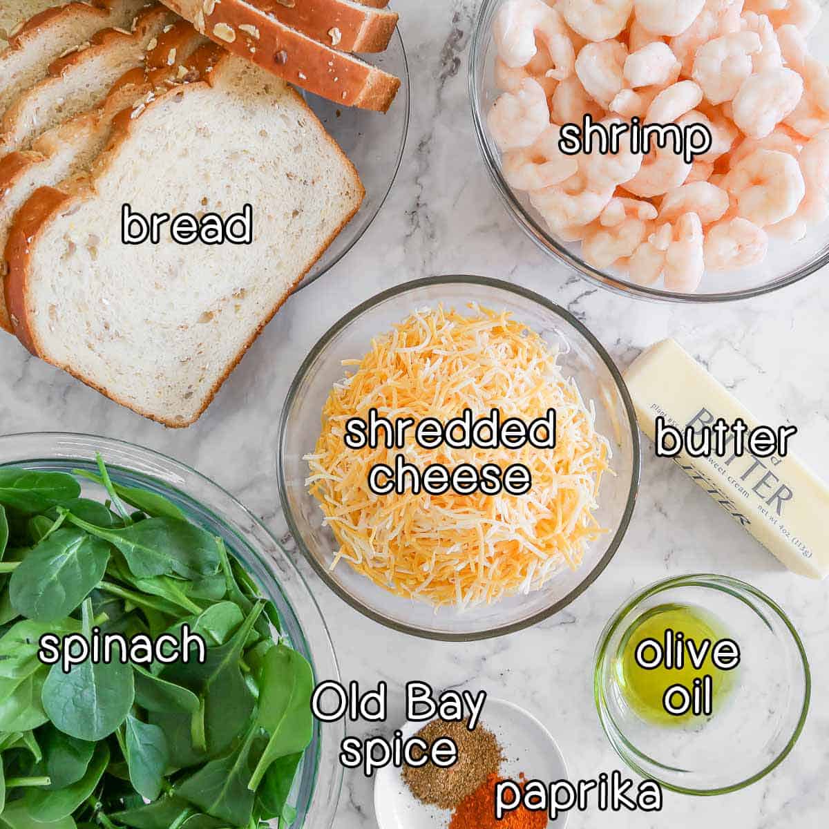 Overhead shot of ingredients- bread, shrimp, shredded cheese, spinach, butter, olive oil, old bay spice, and paprika.