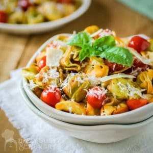 Side angled shot of tortellini garnished with basil and parmesan cheese in a white bowl nestled in another white bowl on a white cloth on a wooden table.